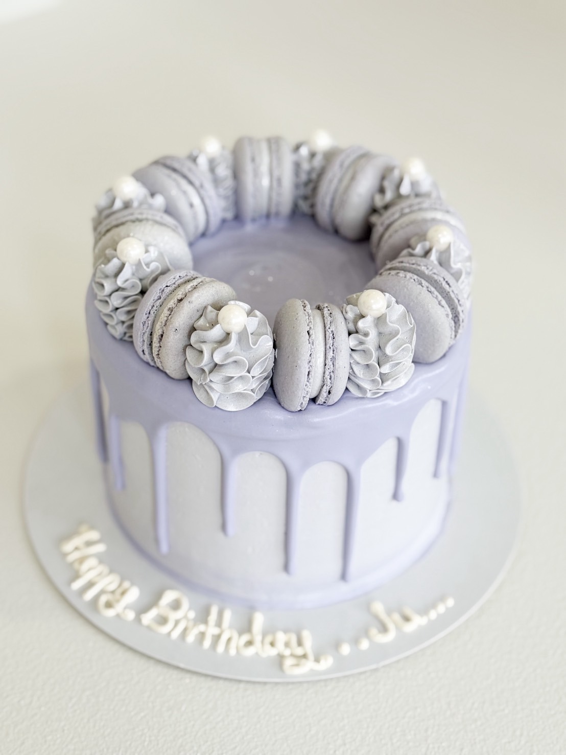 Elegant lavender drip cake topped with matching macarons, whipped cream swirls, and edible pearl accents on a round silver base.