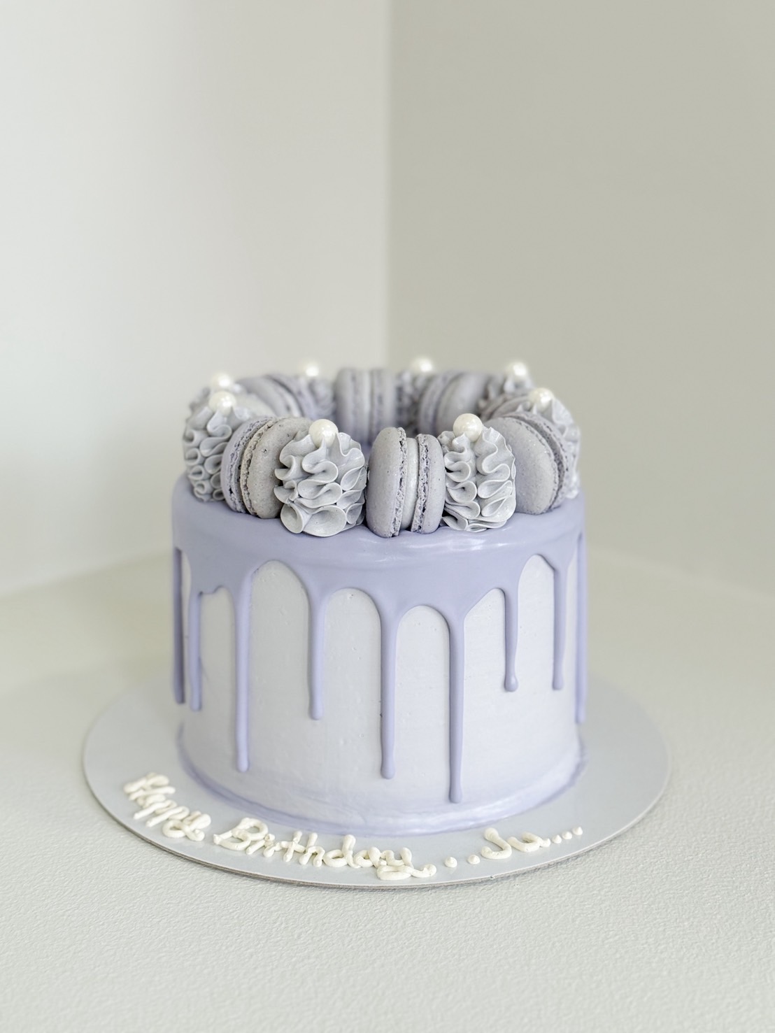 Elegant lavender drip cake topped with matching macarons, whipped cream swirls, and edible pearl accents on a round silver base.