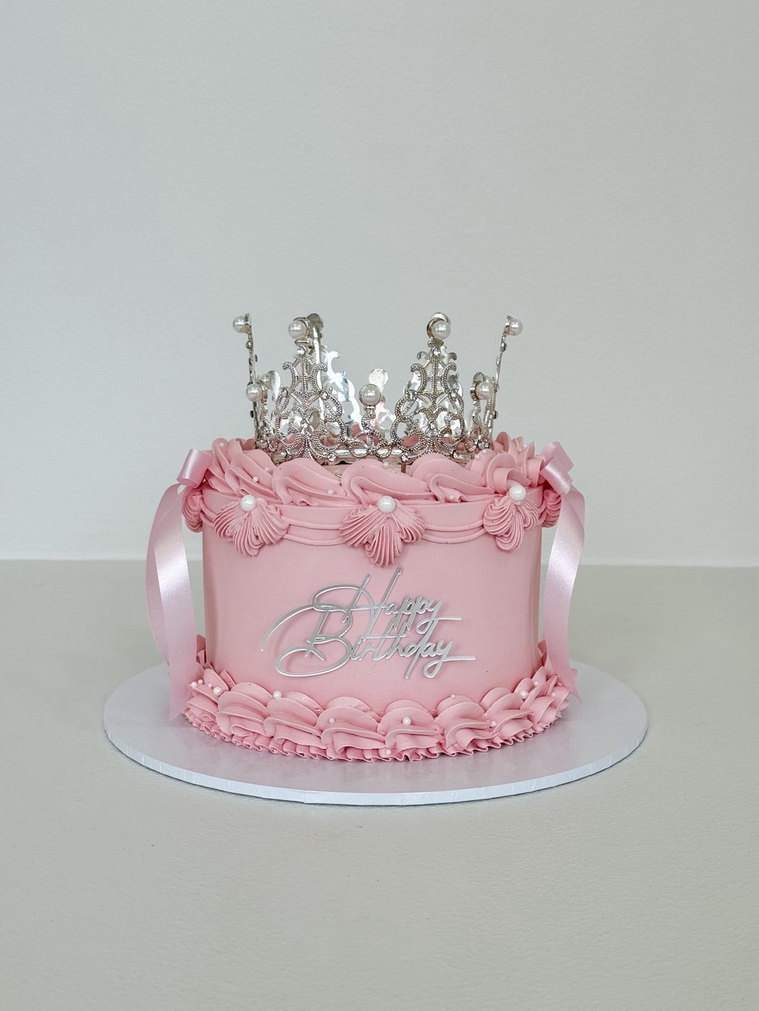 Front view of a pink birthday cake with vintage piping, edible pearls, and silver crown topper