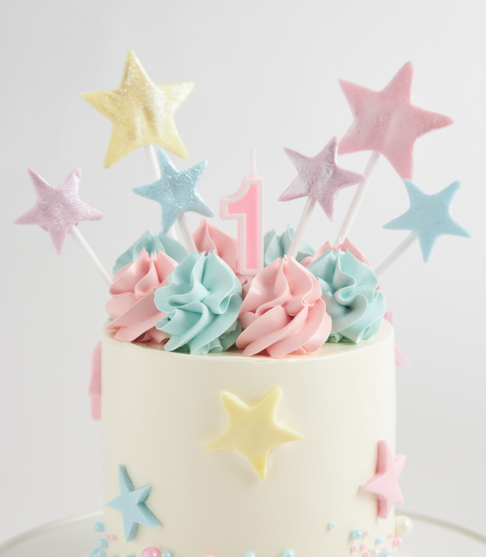 White cake decorated with pastel star toppers, pink “1” candle, and swirls of pink and blue frosting