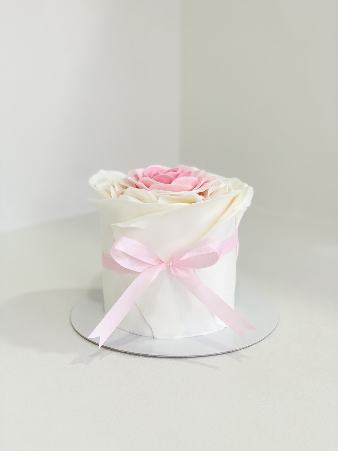 Elegant mini cake with white ruffle icing and a large pink buttercream rose on top, tied with a soft pink ribbon.