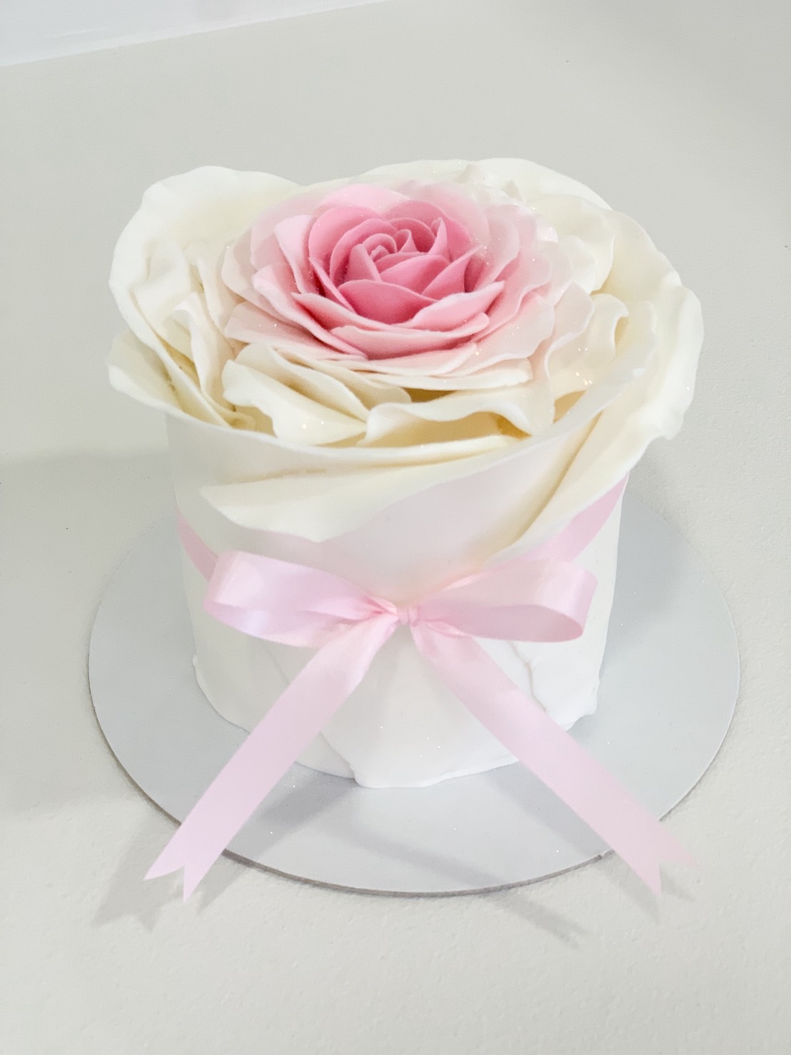 Elegant mini cake with white ruffle icing and a large pink buttercream rose on top, tied with a soft pink ribbon.