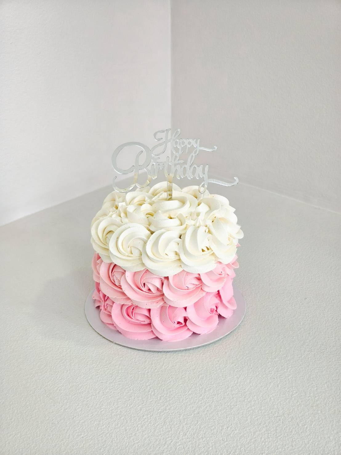 Elegant birthday cake decorated with buttercream rosettes in ombre pink and white, topped with a silver “Happy Birthday” topper.