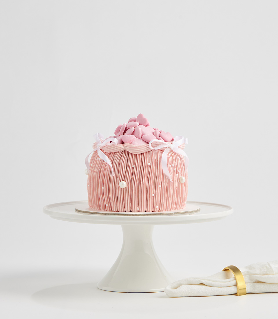 Pink dome-shaped cake topped with fondant hearts, white bows, and pearl decorations