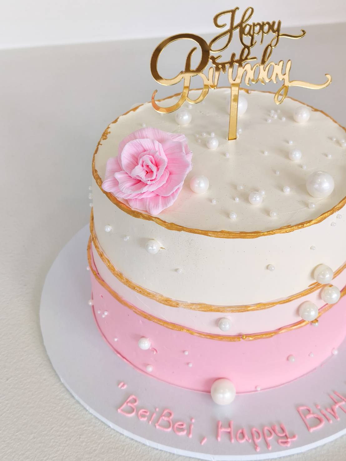 Two-tier birthday cake in white and pink, decorated with edible pearls, golden lines, pink flower, and gold Happy Birthday topper.