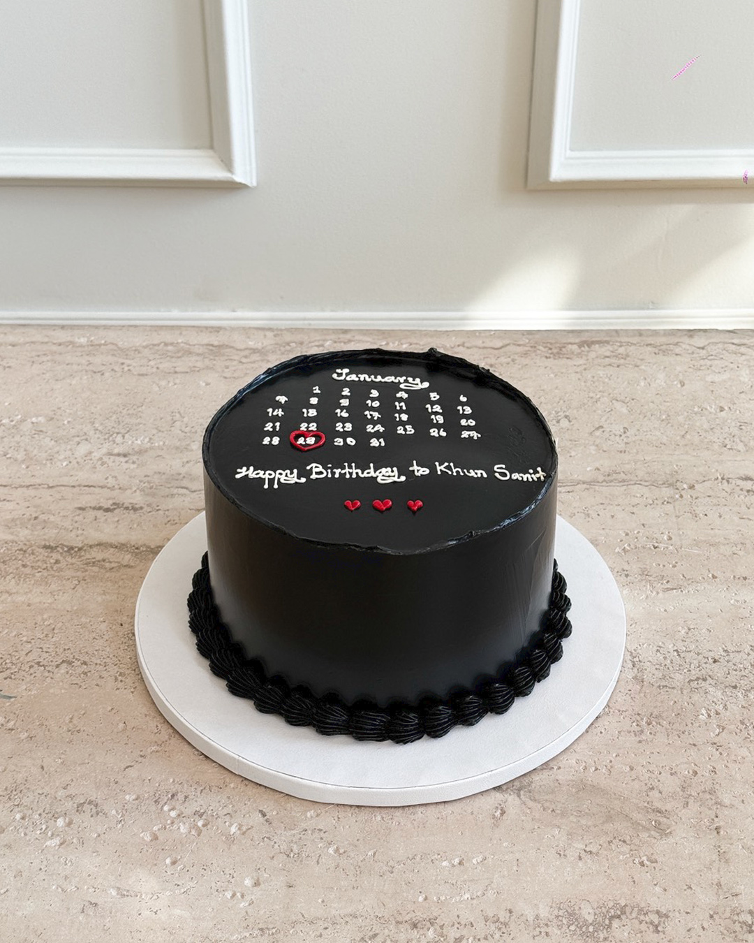 A round black Anniversary Calendar Cake features a white January calendar on top, highlighting the 22nd with a red heart. Decorated with “Happy Birthday to Khan San!” and small red hearts, it sits neatly on a white cake board.