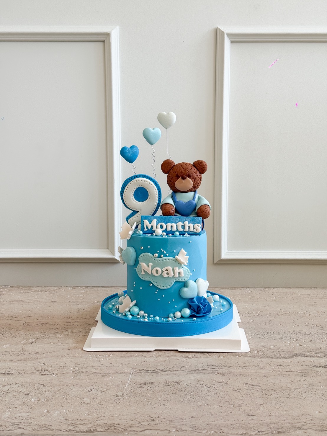 A blue Sweet Bear Baby Cake adorned with a teddy bear, hearts, and “9 Months Noah,” celebrates a 9-month milestone. The cake sits on a white base against a light wall background.