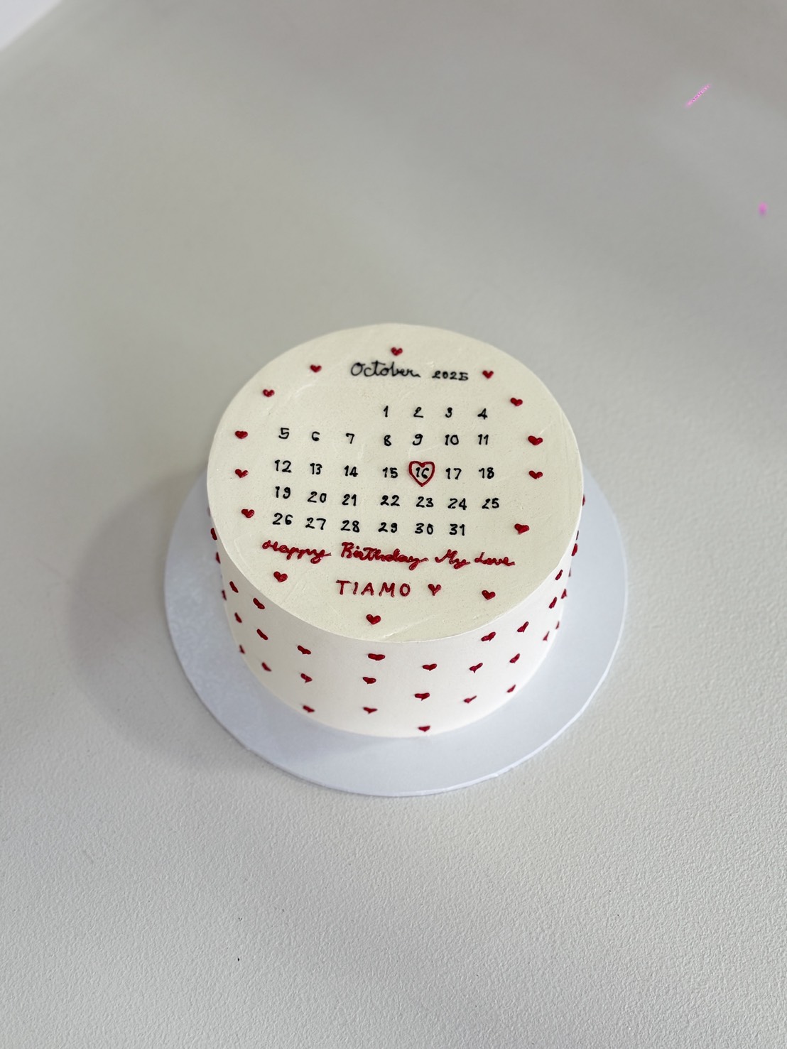 A round white Sweet Calendar Cake decorated with small red hearts and a calendar for October 2023 on top. The 18th is circled with a heart, and 