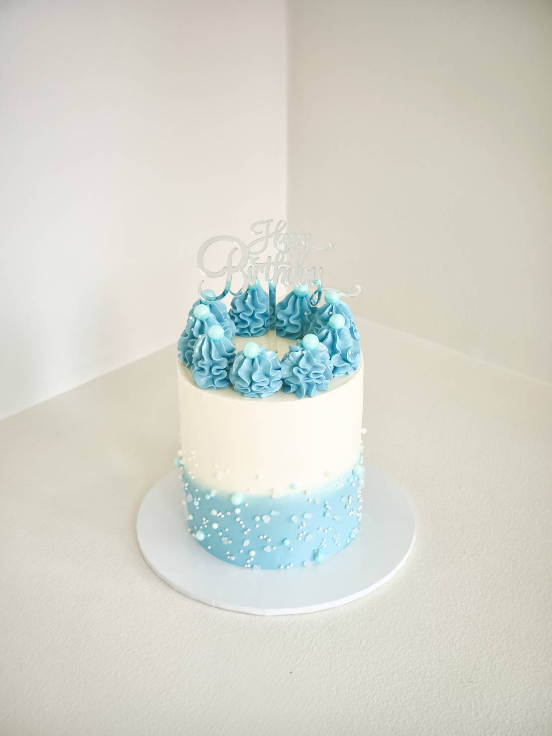 Tall blue and white birthday cake decorated with piped blue cream swirls, edible pearls, and a clear Happy Birthday topper.