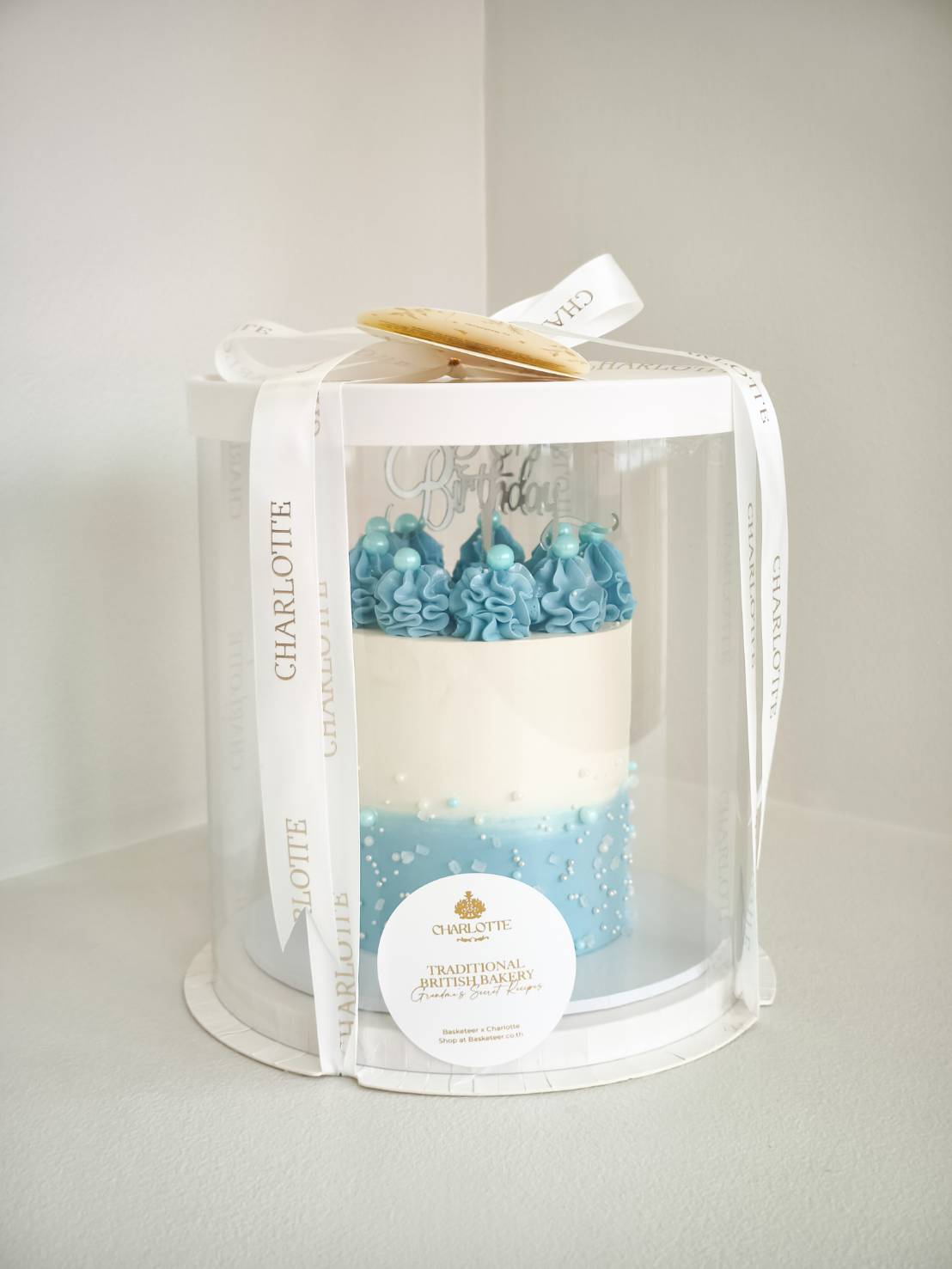 Tall blue and white birthday cake in a transparent luxury gift box with white ribbon and a Happy Birthday topper.