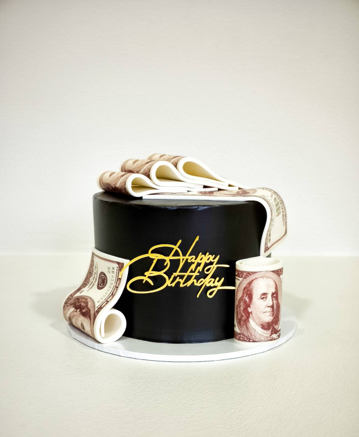 Black birthday cake decorated with edible US dollar bill designs and “Happy Birthday” topper.
