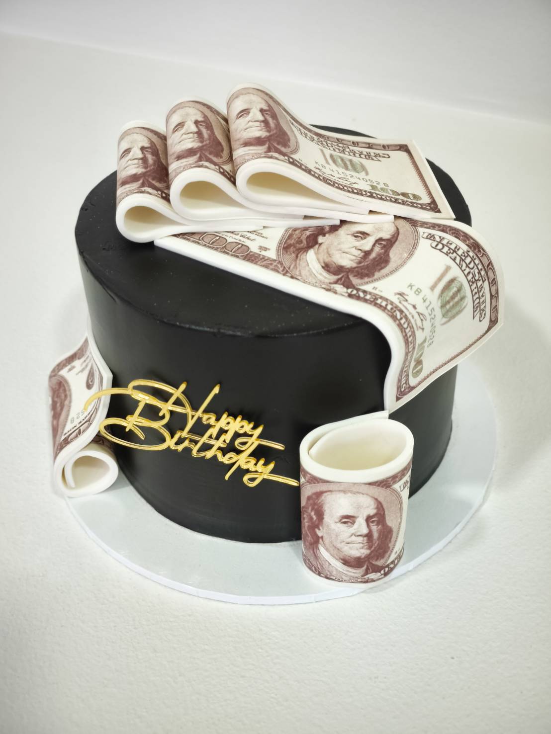 Black cake with edible US dollar bills and golden “Happy Birthday” topper.