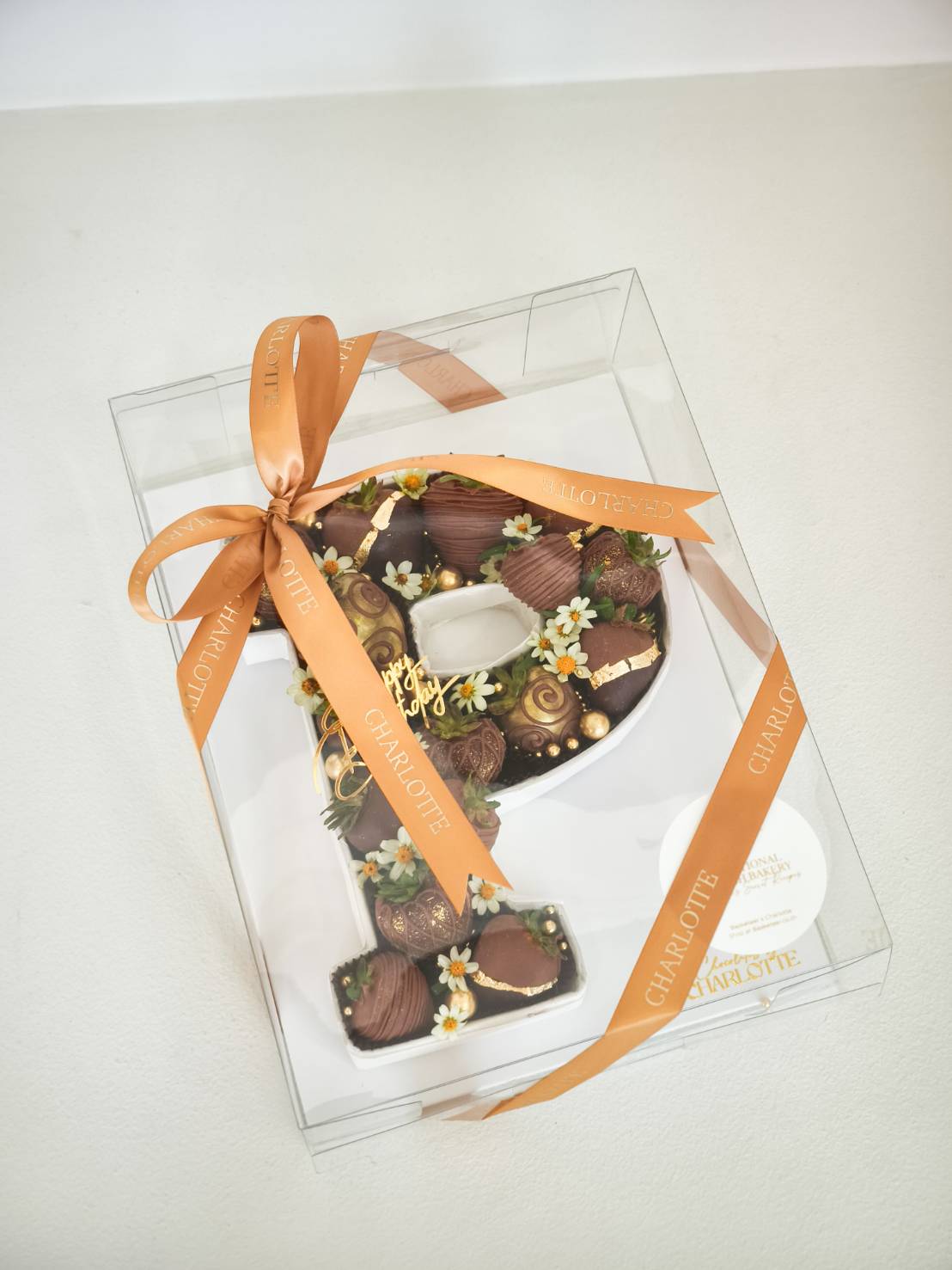 A number 7 chocolate cake, topped with flowers, chocolates, and a Happy Birthday sign, presented in a clear box with an orange Charlotte ribbon.