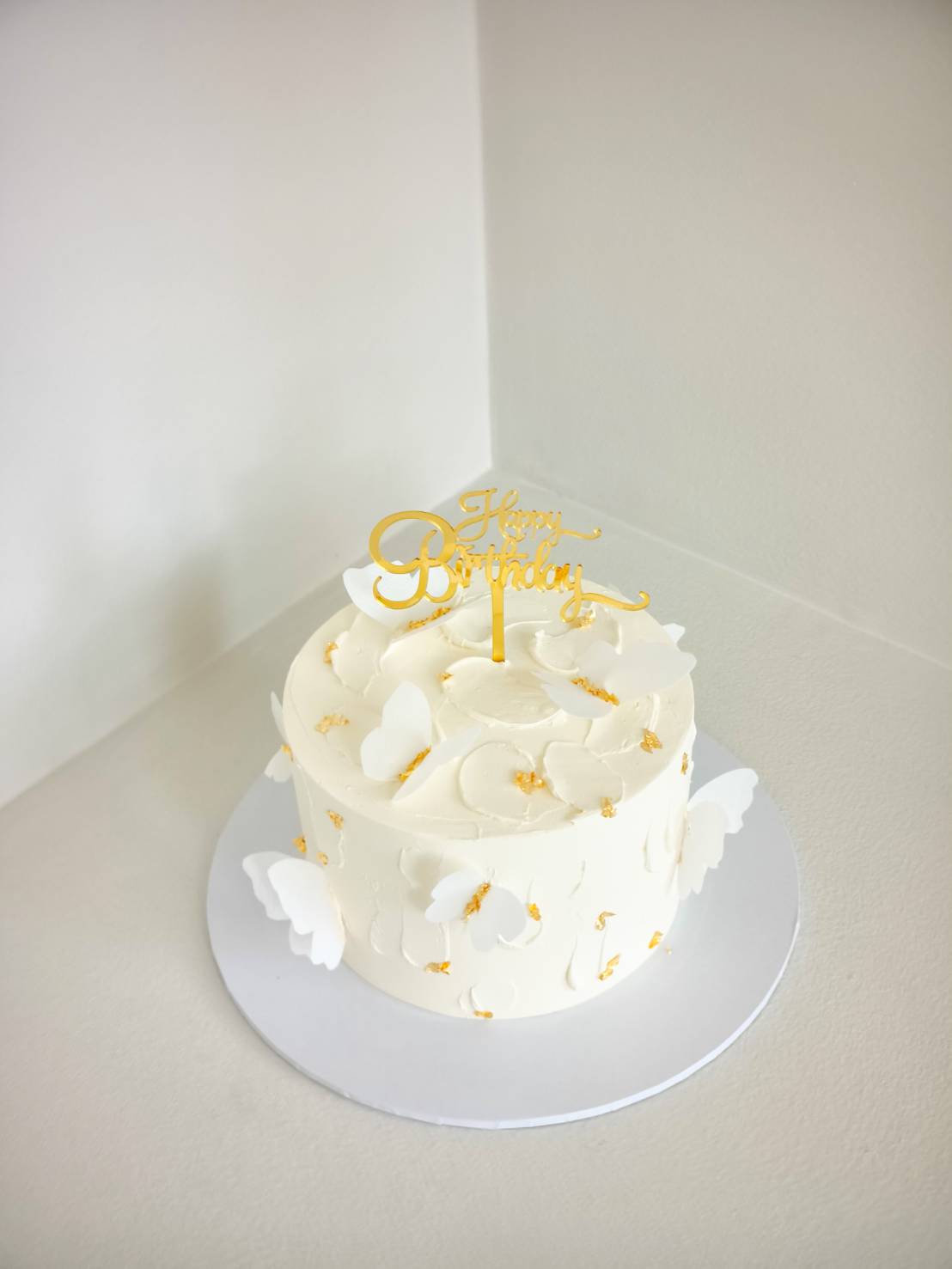 Elegant white birthday cake decorated with delicate edible butterflies and gold accents, topped with a Happy Birthday sign.