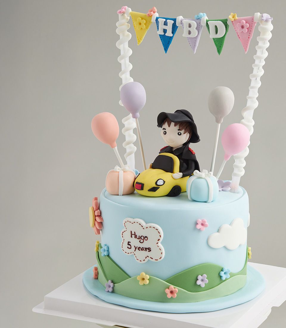 Light blue birthday cake with fondant boy in a yellow car, balloons, and “HBD” banner