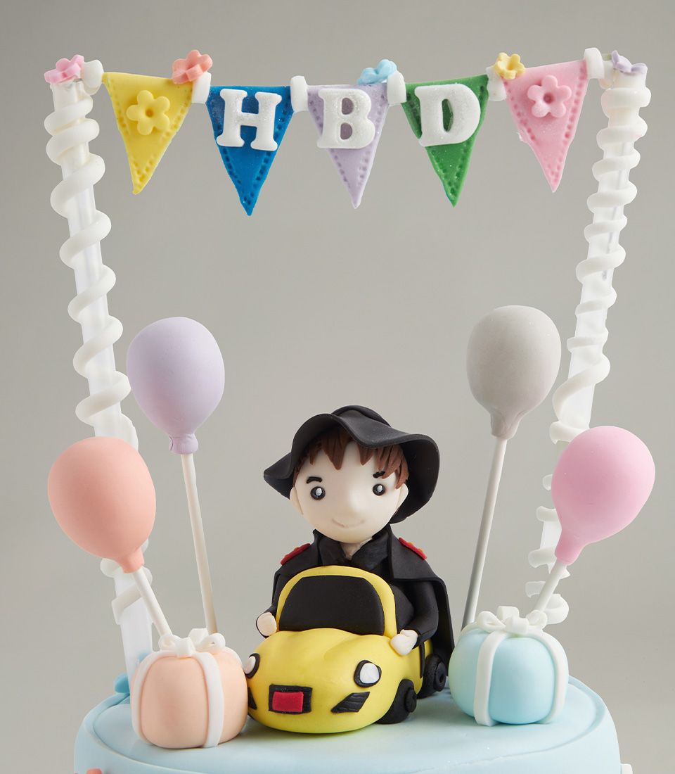 Close-up of fondant boy with black hat sitting in a yellow car surrounded by pastel balloons