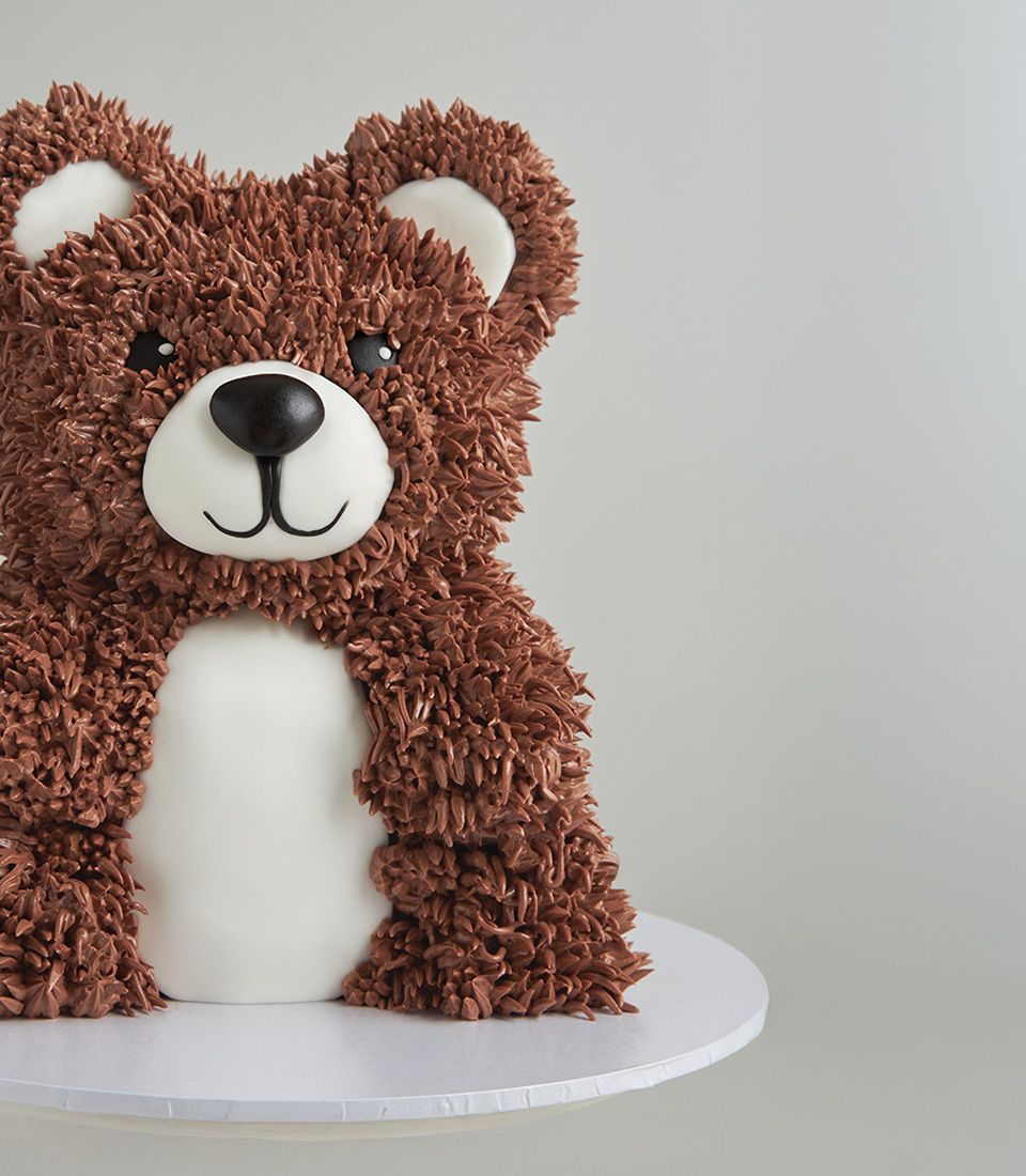 Close-up view of a 3D teddy bear cake made with fondant icing and piped chocolate buttercream fur, featuring a cute white belly and smile.
