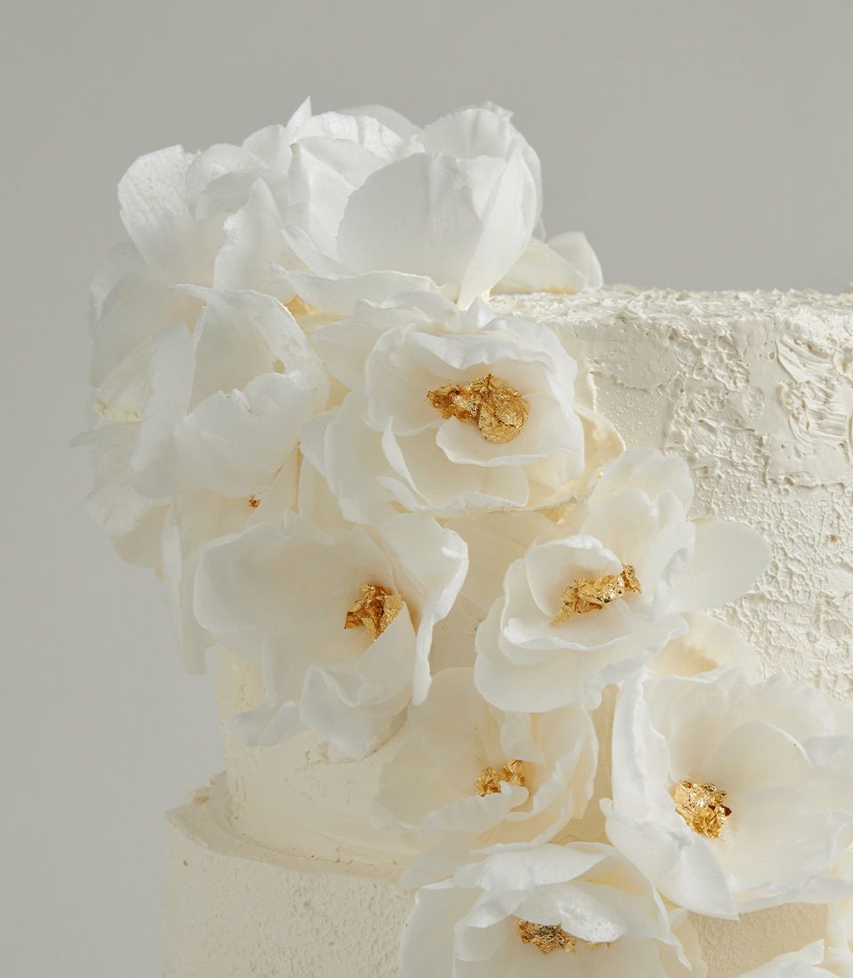 Close-up of a tiered cream cake decorated with delicate white wafer flowers and edible gold leaf, highlighting texture and elegance.