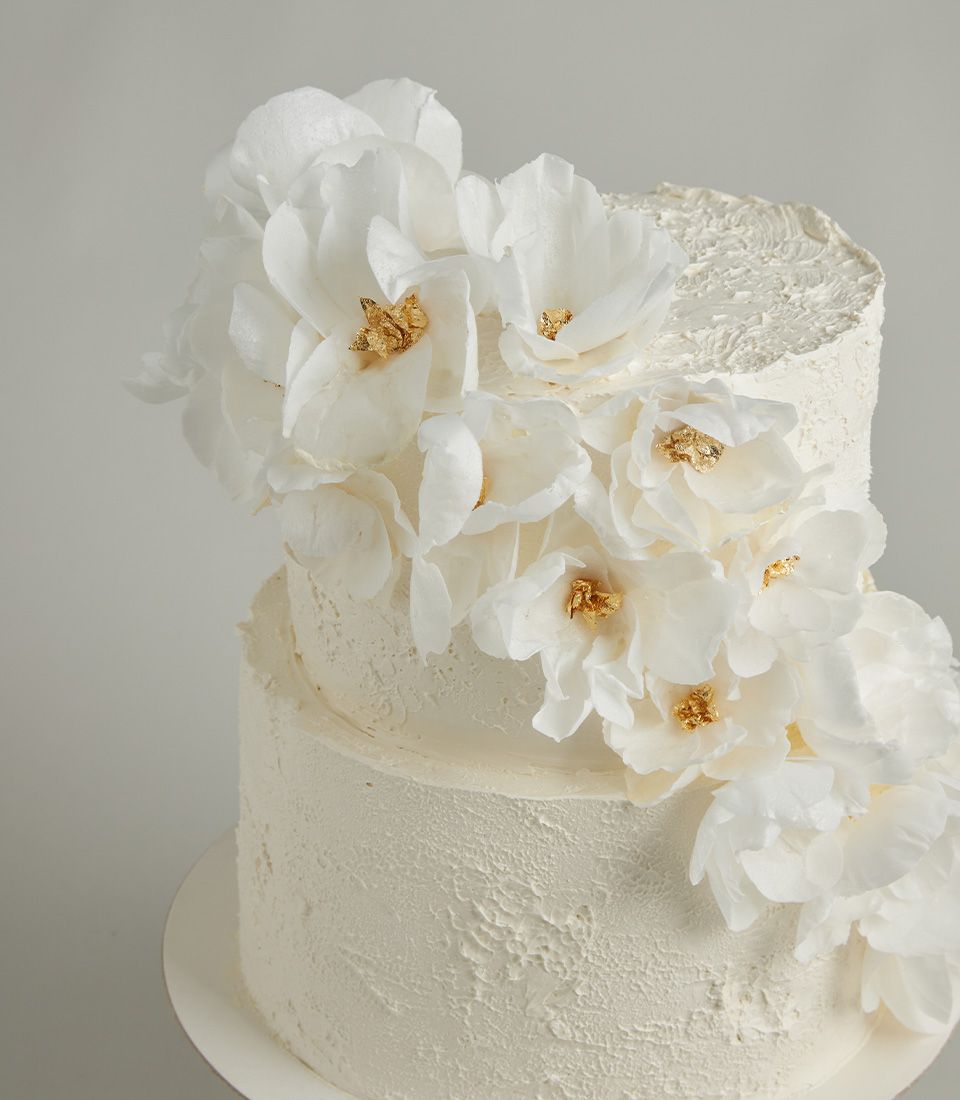 Two-tiered white buttercream cake adorned with cascading white wafer flowers and edible gold leaf for a luxurious and elegant design.