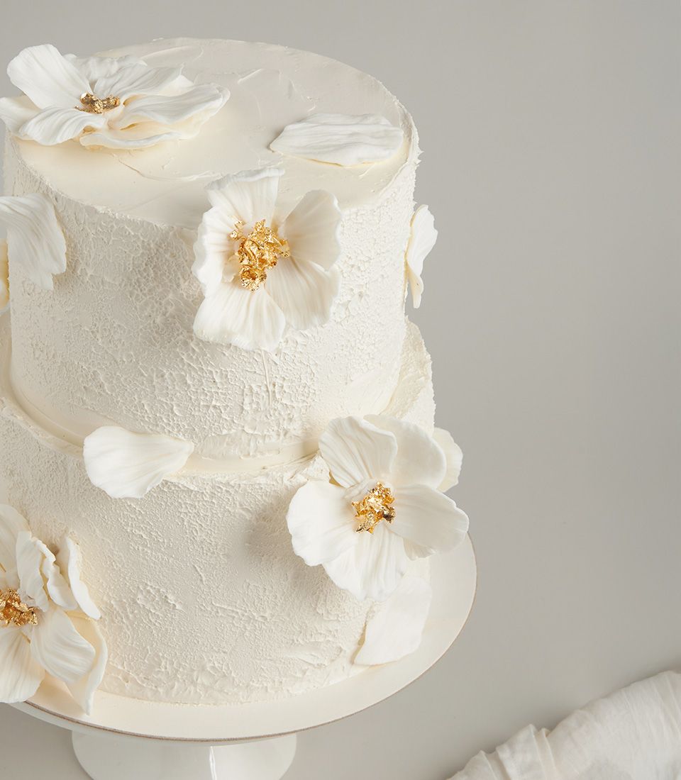 Two-tier white fondant wedding cake with textured buttercream finish, adorned with sculpted white flowers and gold leaf centres.