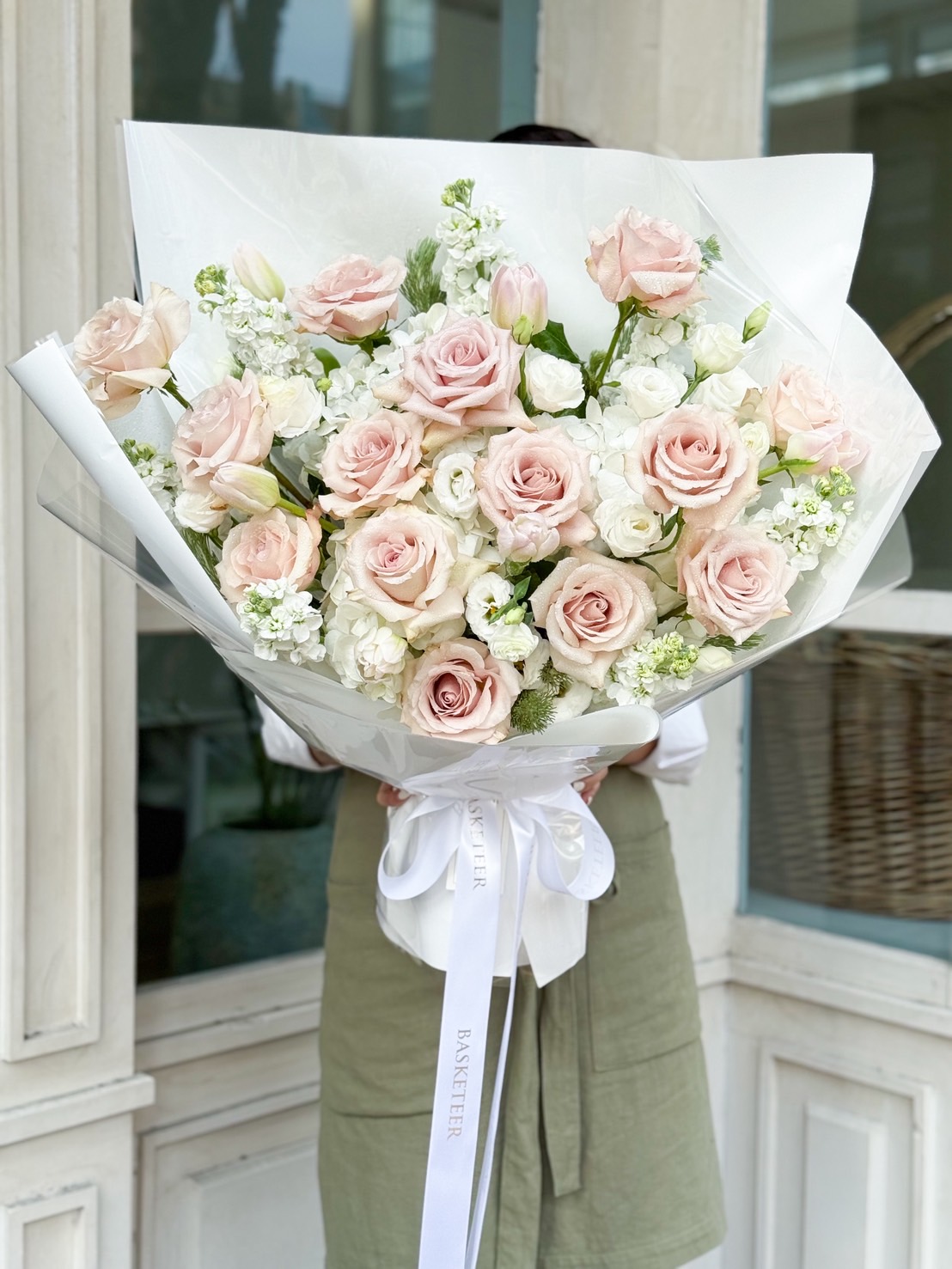 Luxury bouquet of blush pink roses, white lisianthus, hydrangeas, and tulips, wrapped in white paper with satin Basketeer ribbon.