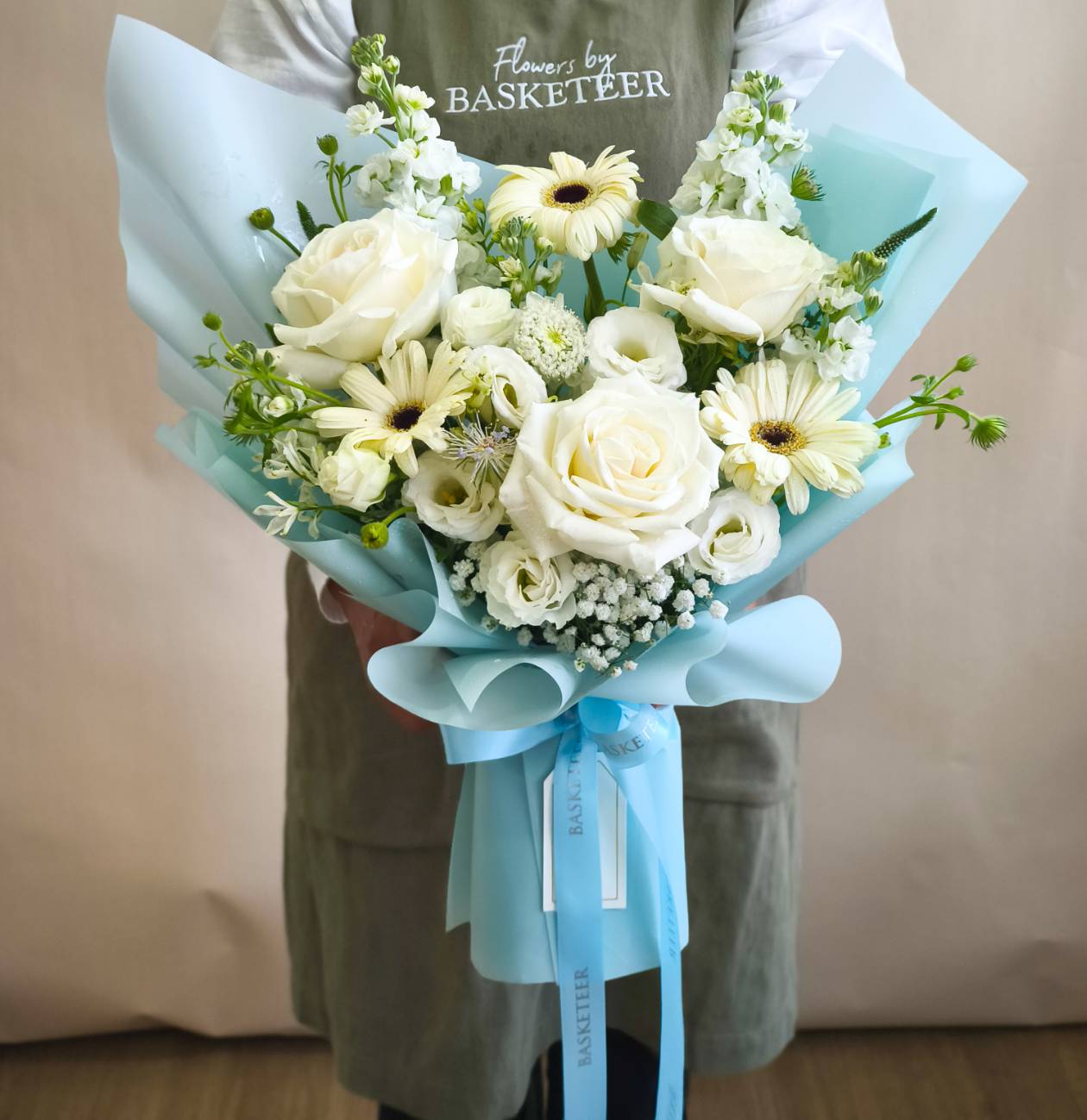 Elegant bouquet with white roses, cream gerberas, lisianthus, gypsophila, and green accents, wrapped in pastel blue paper with Basketeer ribbon.