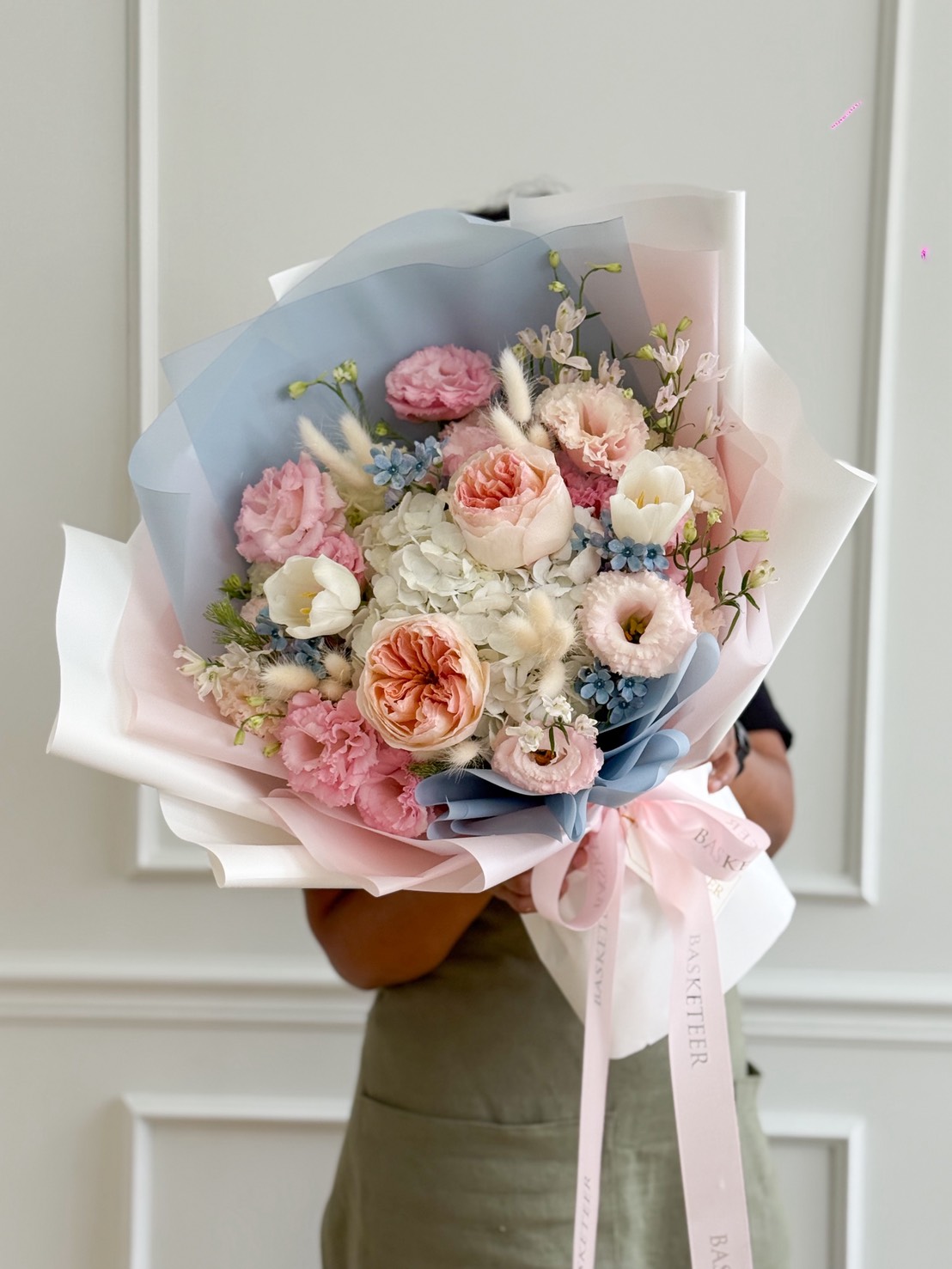 Pastel bouquet with roses, hydrangeas, carnations, lisianthus, tulips, and bunny tail grass wrapped in pink and blue layered paper with ribbon