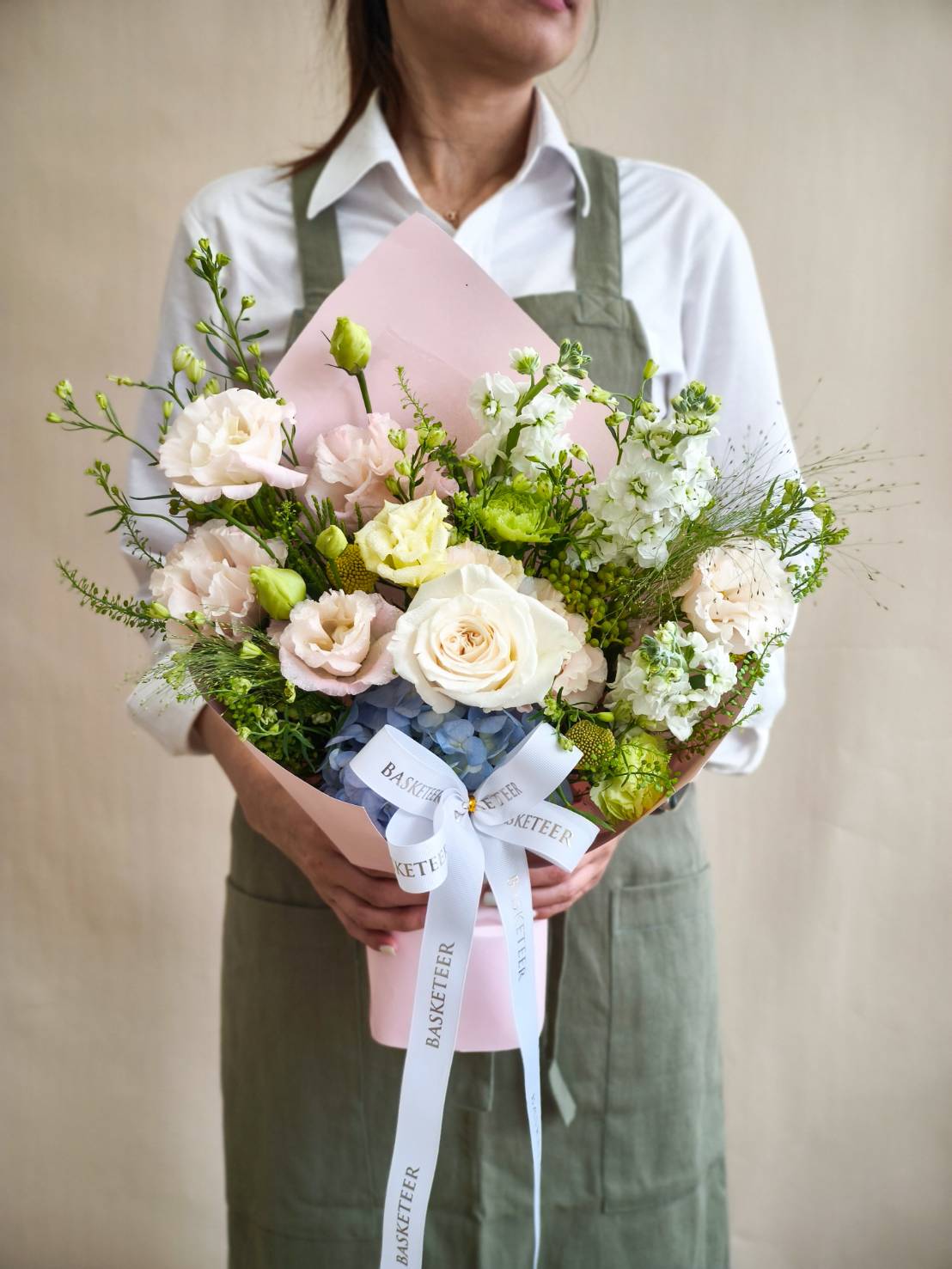 Elegant pastel bouquet with cream roses, light lisianthus, blue hydrangeas, and soft greenery wrapped in pink paper with ribbon.