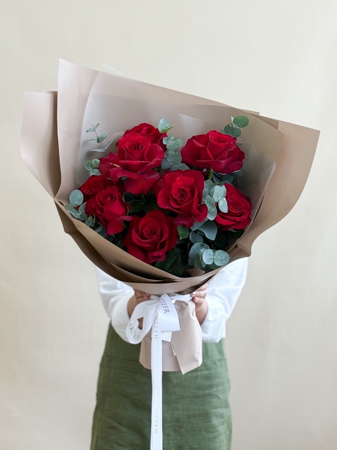 Elegant bouquet of fresh red roses with eucalyptus, wrapped in beige paper and tied with Basketeer ribbon.