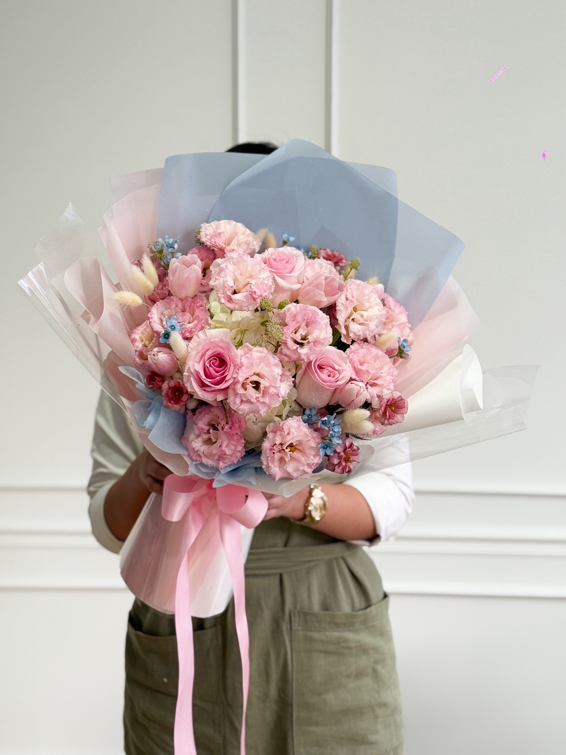 A person in a white shirt and green apron holds a large Pastel Charm Flower Bouquet wrapped in pastel blue and white paper, tied with a pink ribbon. The bouquet covers the person's face.