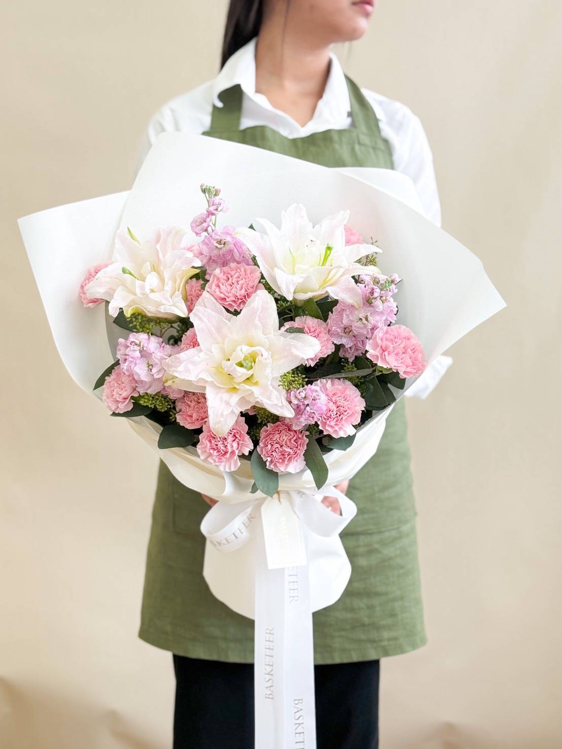 Elegant bouquet of pink carnations, white lilies, and pastel flowers wrapped in white paper with a satin ribbon by Basketeer