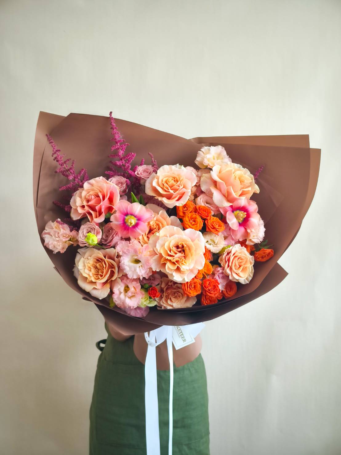 Elegant bouquet of peach, orange, and pink roses with astilbe and mixed blooms, wrapped in rich brown paper.