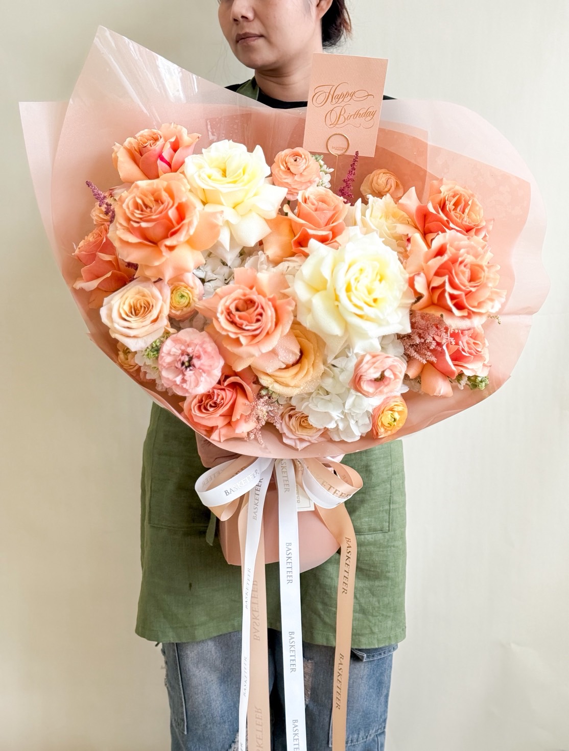 Large bouquet of peach, coral, and yellow roses with white hydrangeas and pastel flowers, wrapped in peach paper with satin ribbons.