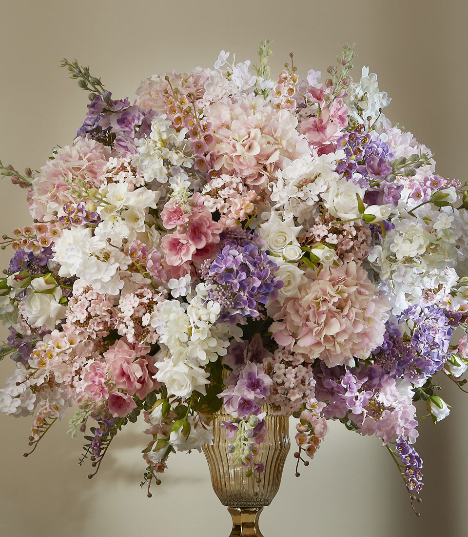 A large, elegant bouquet of pastel flowers, including pink, purple, and white blooms, arranged in a tall glass vase against a neutral background.