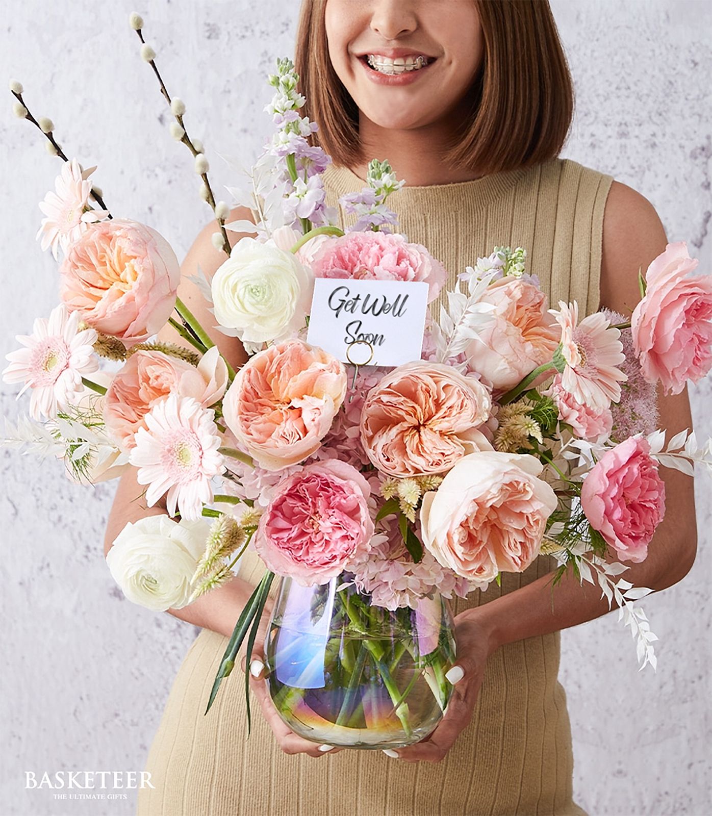 A woman in a beige sleeveless dress smiles and holds a glass vase filled with pink and peach flowers, with a card reading “Get Well Soon” nestled among the blooms.