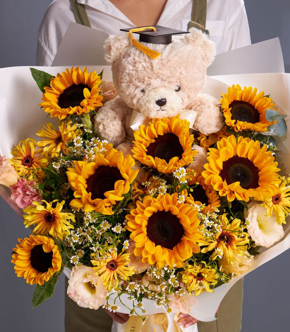 A person holds a large bouquet of sunflowers and other flowers, with a cream-colored teddy bear wearing a graduation cap nestled in the center.