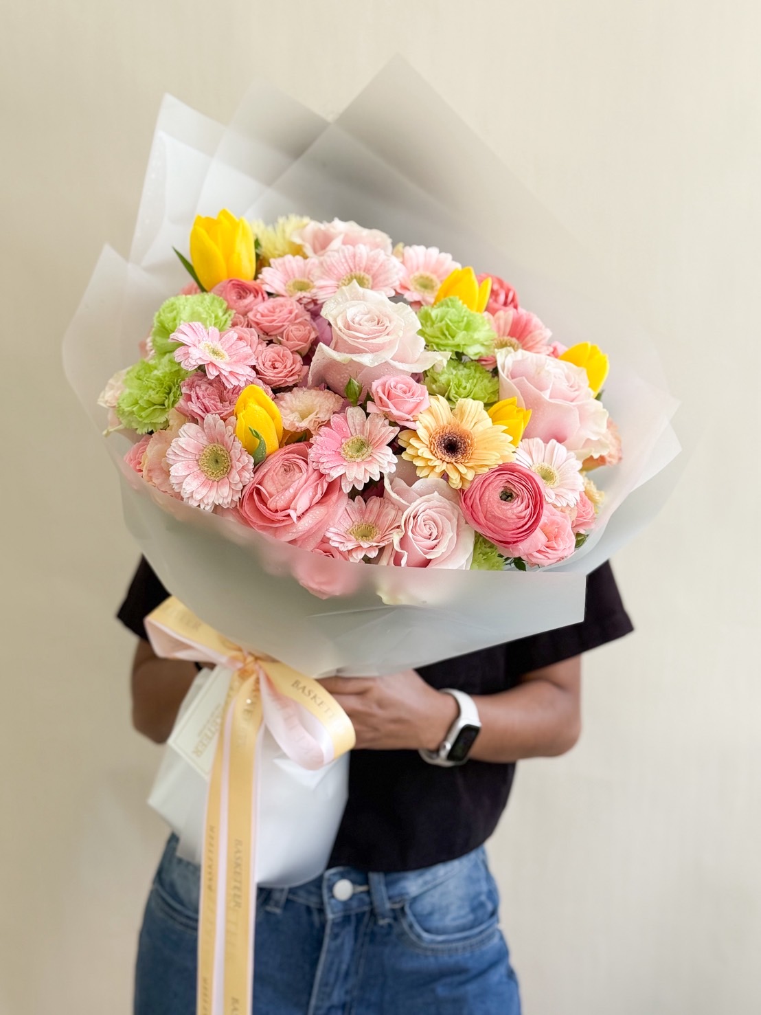 Luxury bouquet with pastel roses, yellow tulips, pink gerberas, green carnations, and ranunculus, wrapped in white paper.