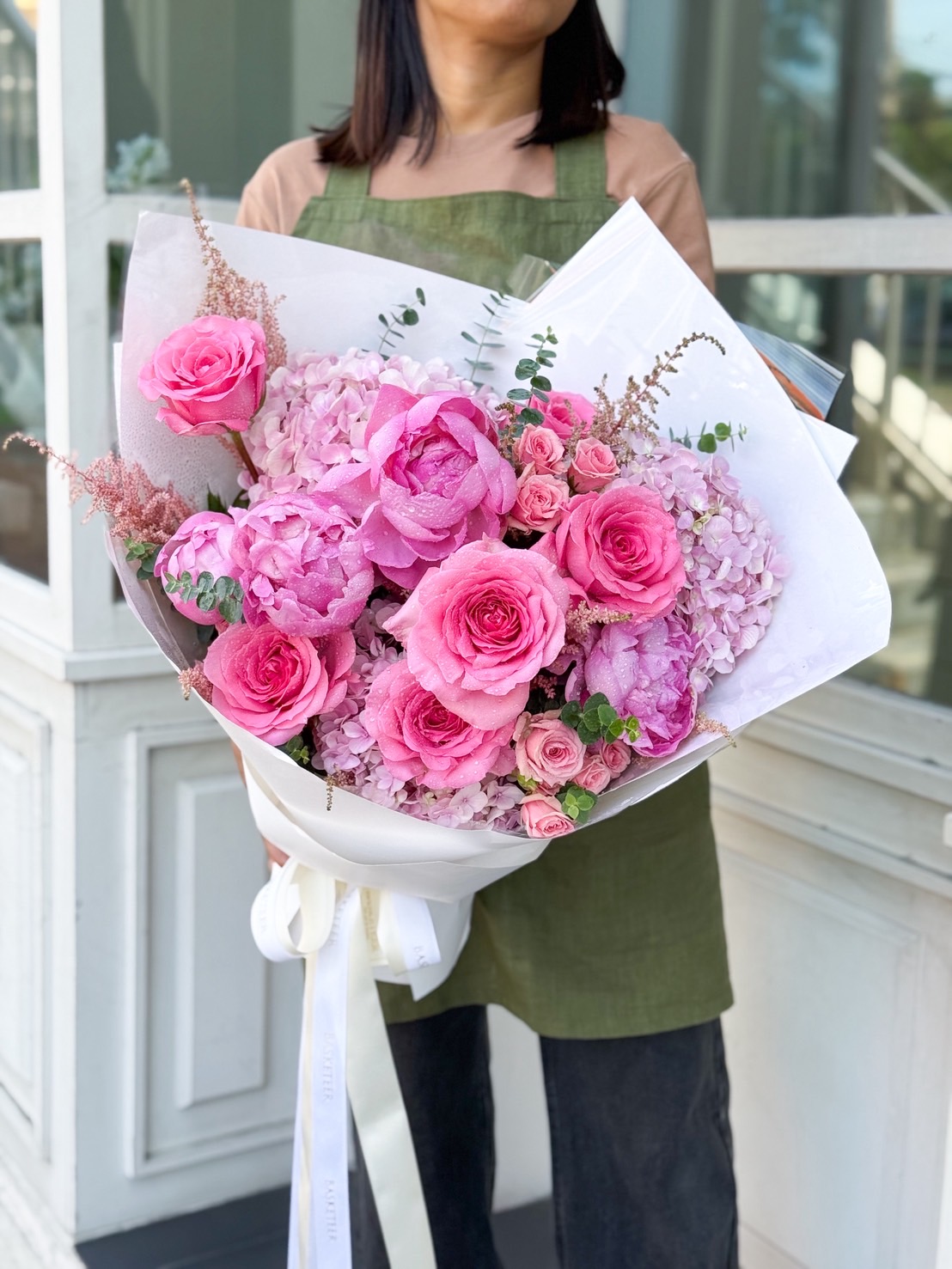 Elegant bouquet of pink peonies, roses, and hydrangeas wrapped in white paper, held by a florist in a green apron.