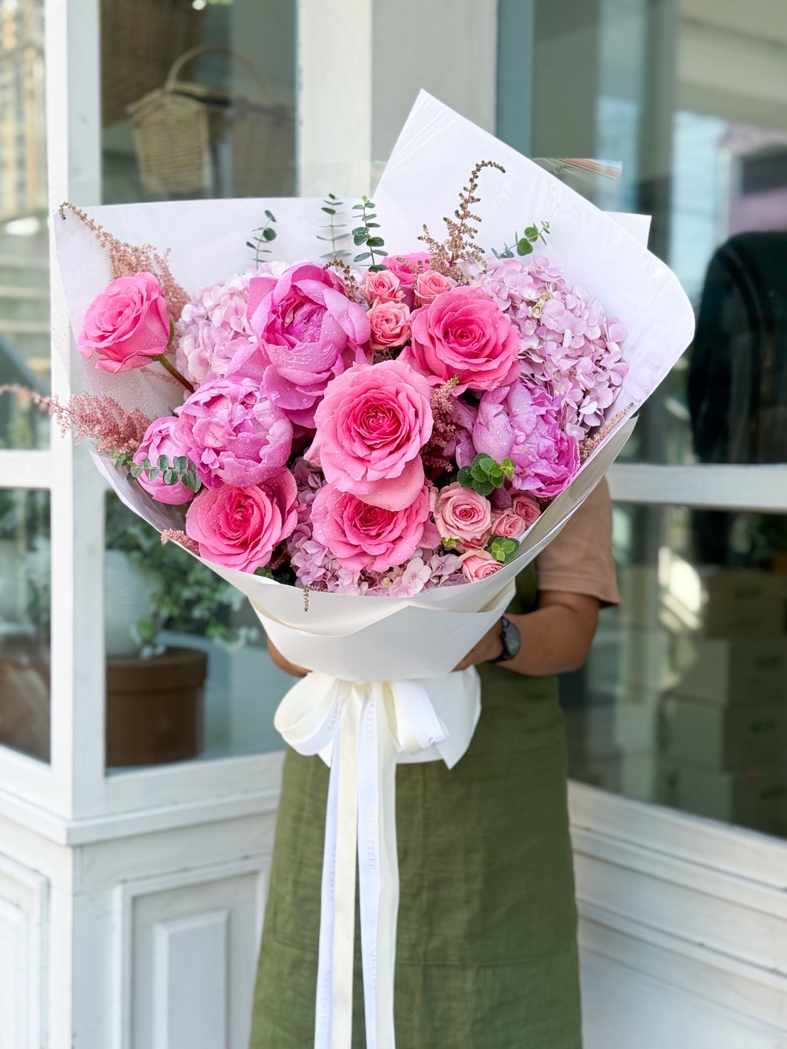Elegant bouquet of pink peonies, roses, and hydrangeas wrapped in white paper, held by a florist in a green apron.