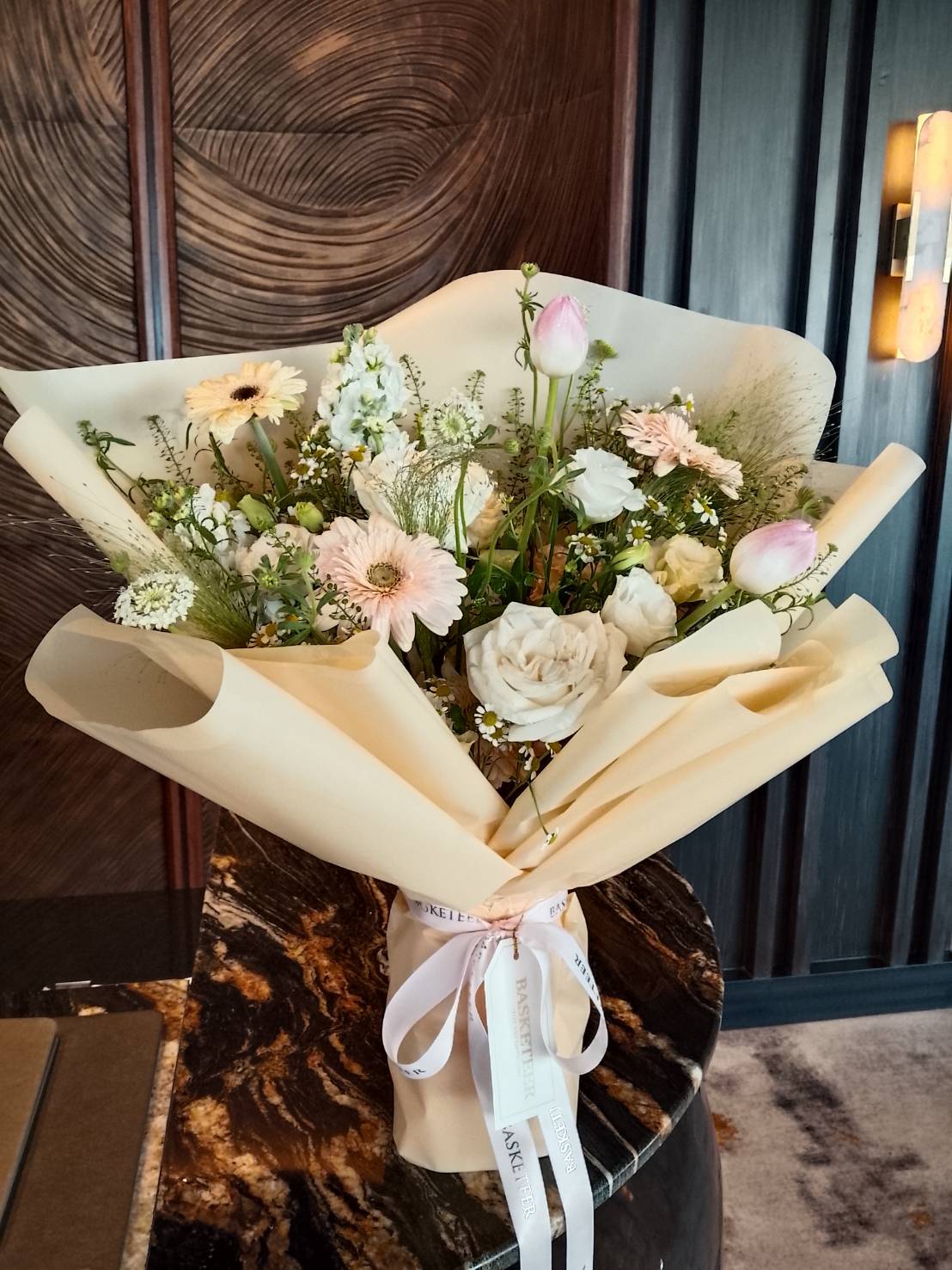 A Charming Mixed Bloom Bouquet of white and pale pink flowers, wrapped in light beige paper and tied with white ribbons, is displayed on a marble-topped table in a modern interior with dark wood panelling.