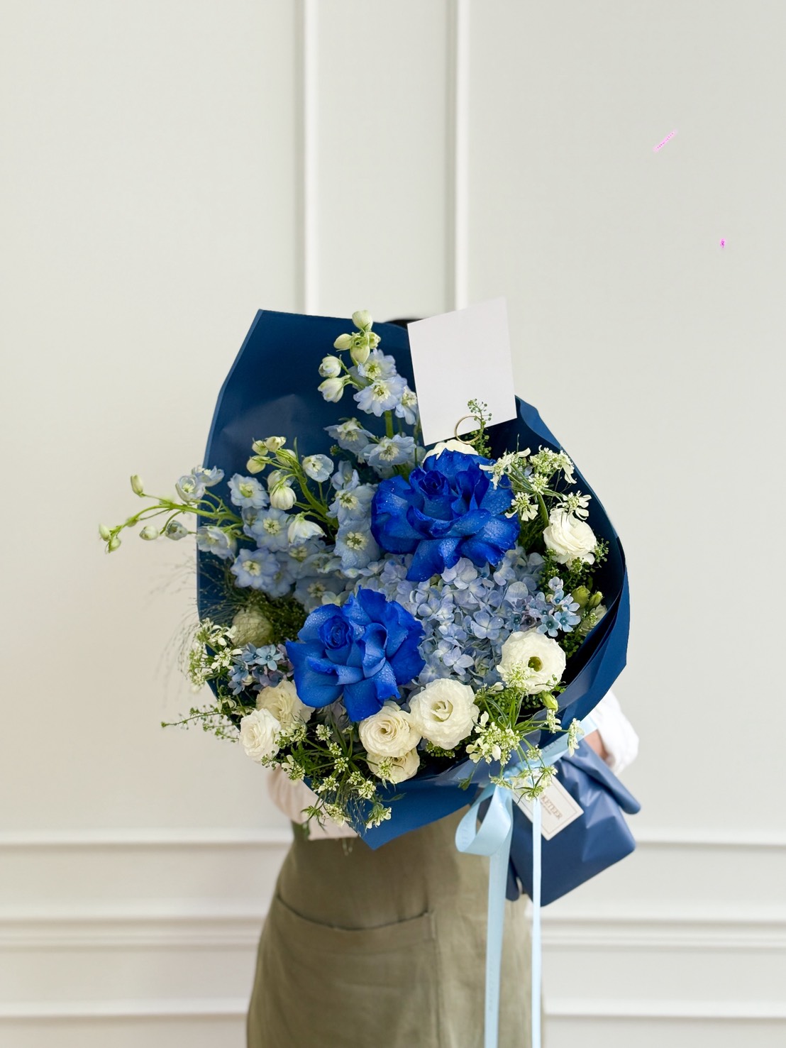 A person holding a stunning Blue Rose & Hydrangea Bouquet wrapped in dark blue paper, with a blank card attached, stands in front of a white wall.