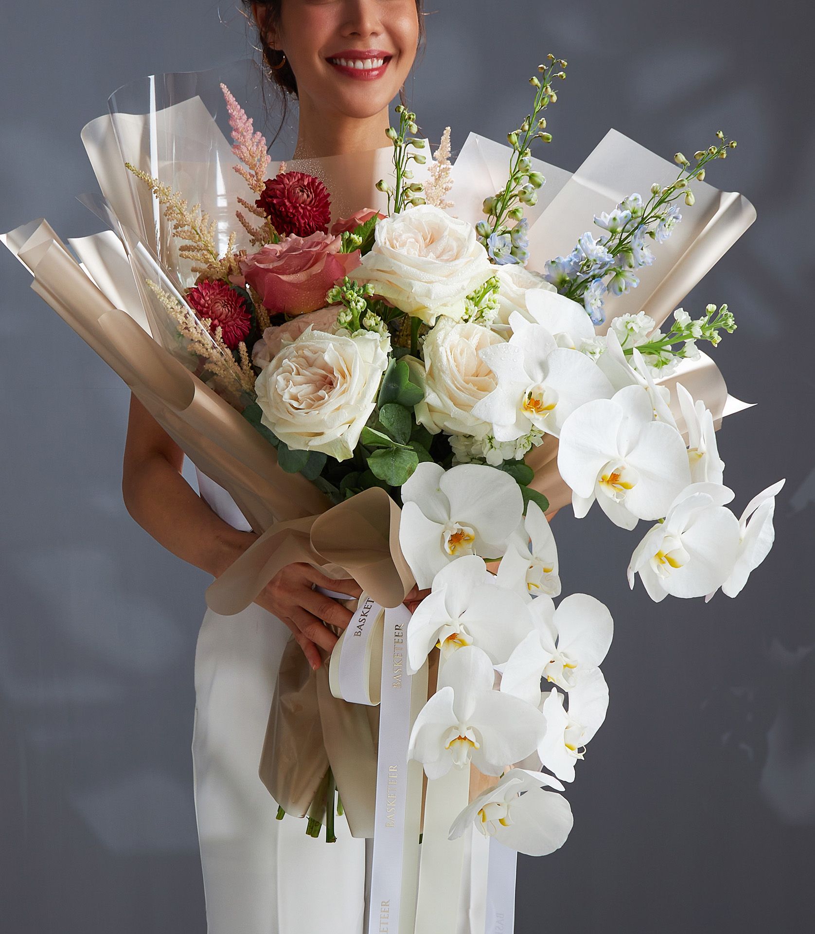 Woman holding luxury bouquet of Moab and O’Hara roses, cascading white orchids, red dahlias, blue delphinium, and astilbe