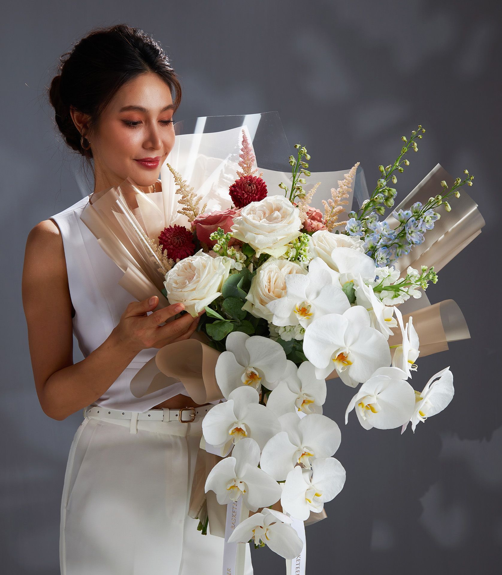 Elegant woman holding a luxury bouquet of white orchids, O’Hara roses, Moab roses, and red dahlias with beige wrapping