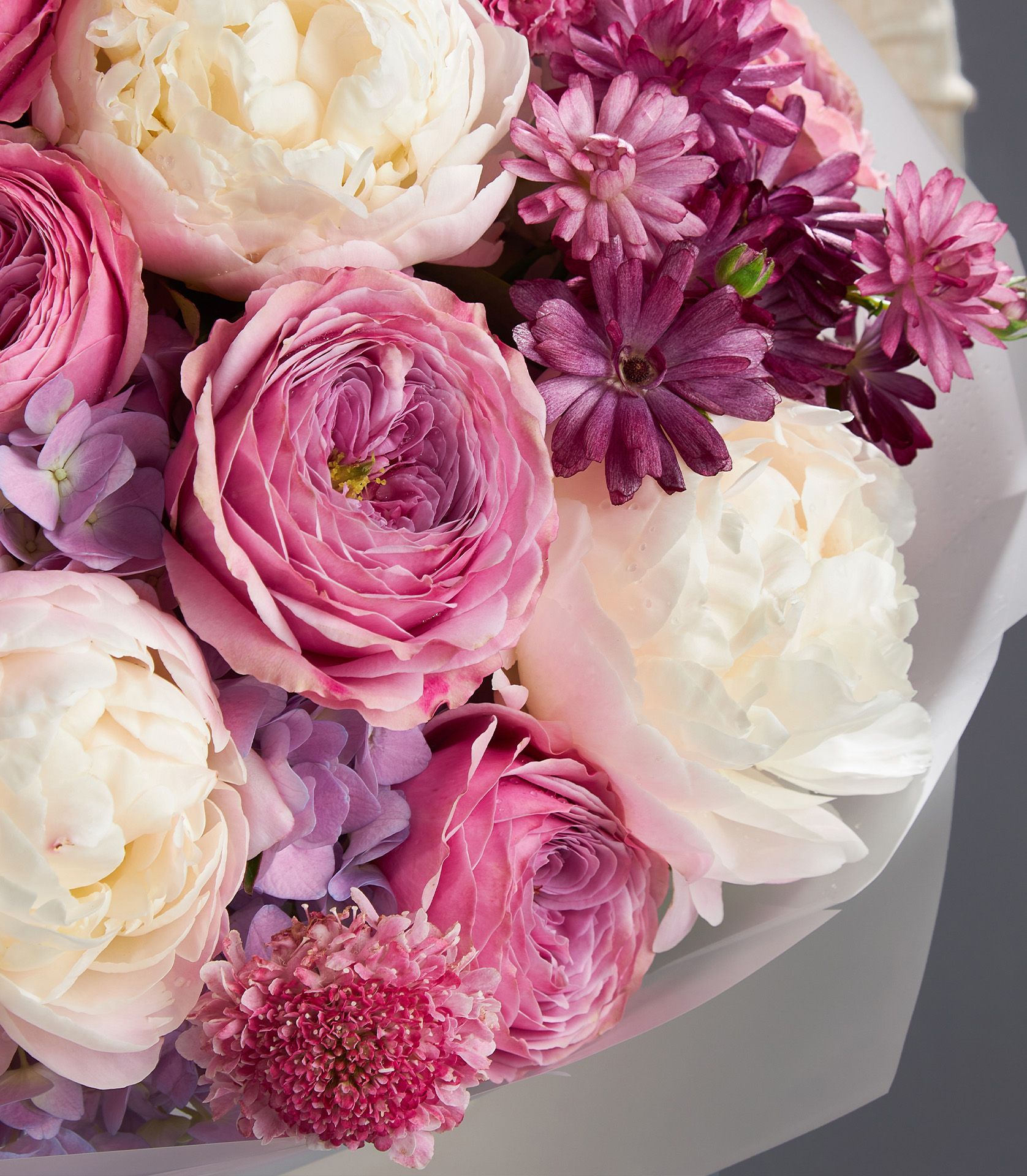 A premium bouquet of white peonies, Peonikiss roses, hydrangeas, butterfly ranunculus, and scabiosa in soft pink and rich purple tones.