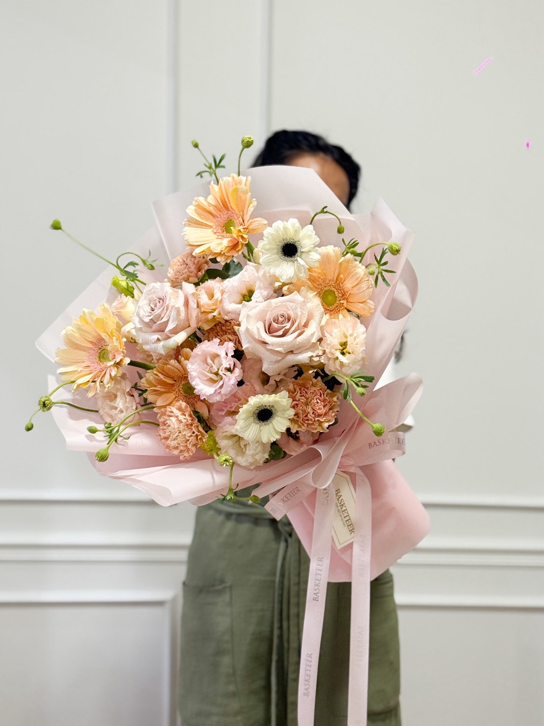 A person holds a large Romantic Blush Mixed Bouquet of pastel pink and peach flowers with light green stems, wrapped in pale pink paper and ribbon, standing against a white wall. The bouquet covers most of the person's face.
