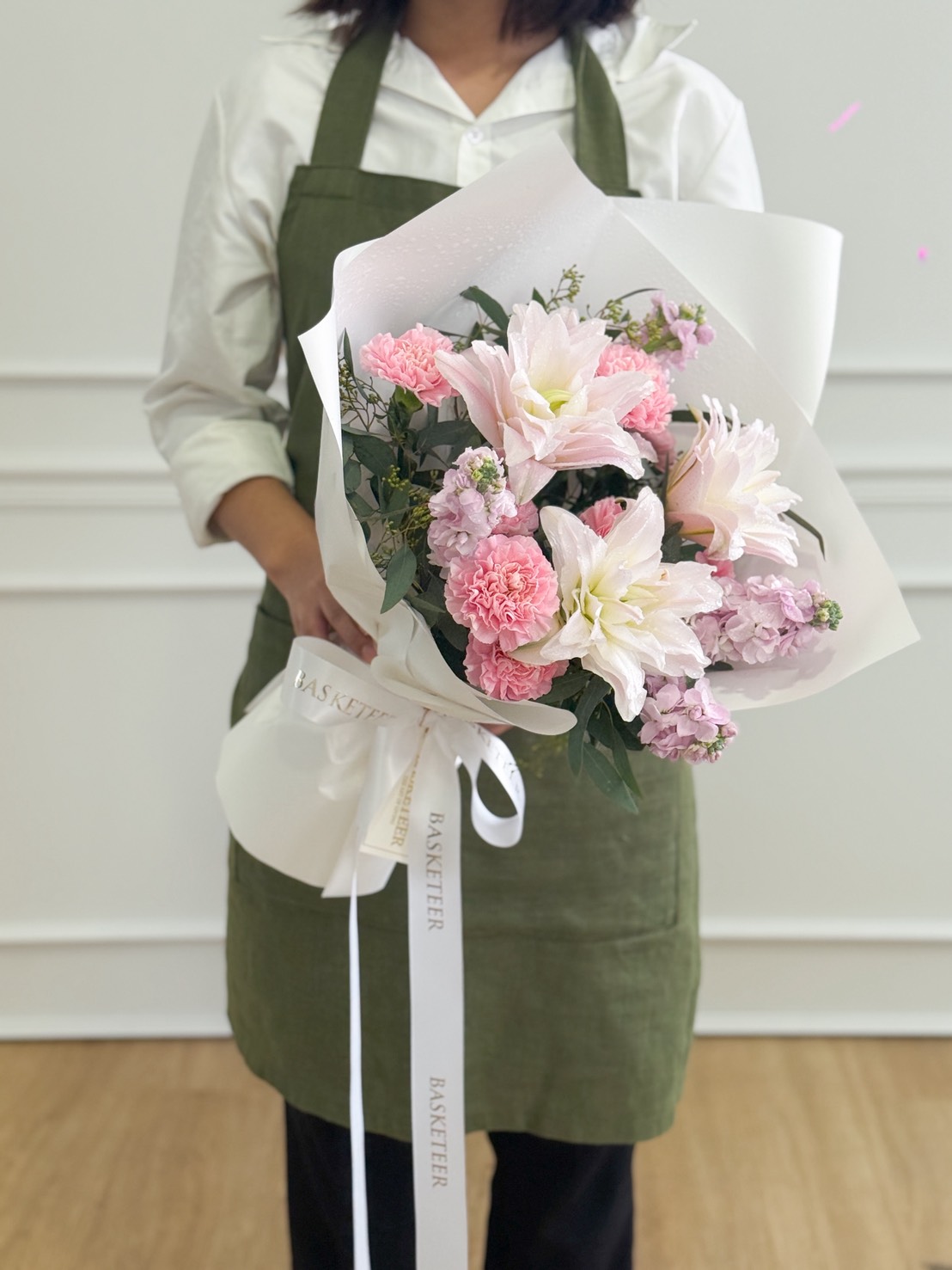 A person wearing a white shirt and green apron holds a Romantic Pink Lily Bouquet wrapped in white paper, tied with a ribbon that reads “BASKETTER.” Pink lilies and white flowers create an elegant, heartfelt arrangement.