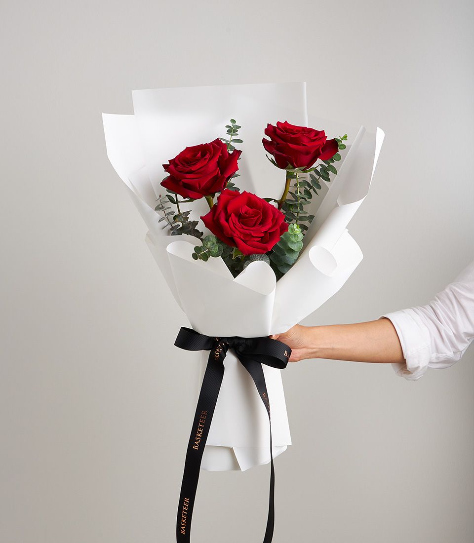 Bouquet of three premium red roses with eucalyptus leaves, wrapped in elegant white paper and tied with a black satin ribbon.