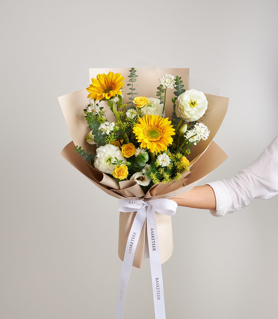 A bright mix of sunflowers, yellow spray roses, ranunculus, chamomile daisies, and eucalyptus in matte beige wrapping.