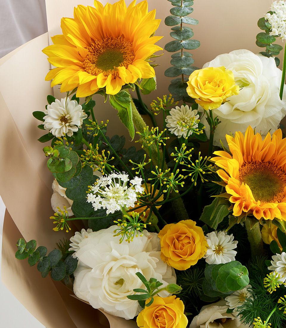 Close-up of a bright bouquet featuring sunflowers, yellow spray roses, white ranunculus, white daisies, and eucalyptus in matte beige wrapping.