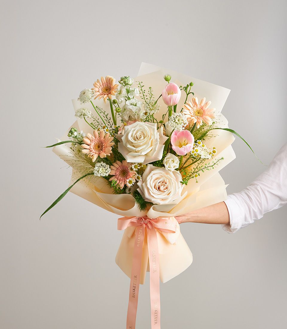 A pastel bouquet of cream roses, peach gerberas, pink tulips, and white blooms with lush greenery, wrapped in soft peach paper and tied with a satin ribbon.